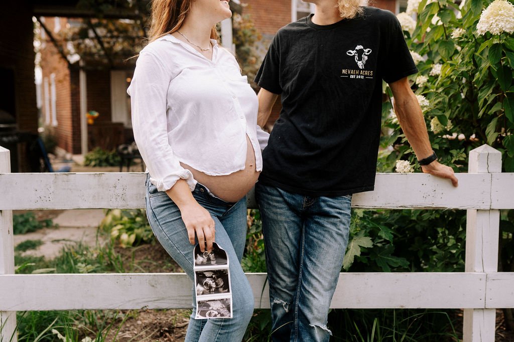 Expectant couple sitting on white wooden fence, woman in white top and man in black shirt