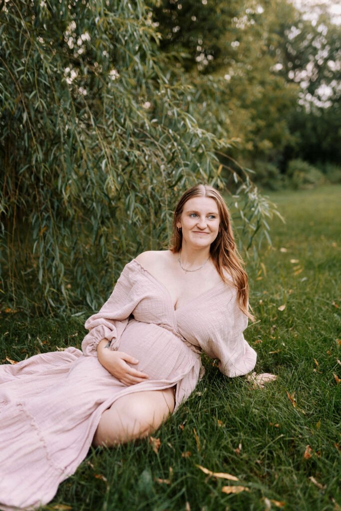 Pregnant woman in flowing blush pink gown sitting on grass under weeping willow tree