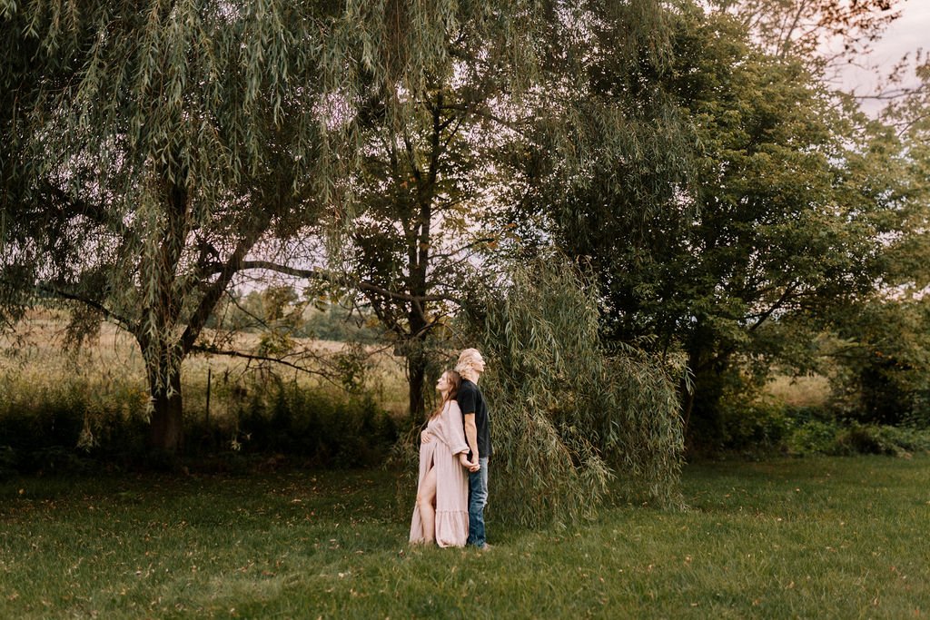 Maternity portrait of woman in beige dress under large willow tree in golden hour light