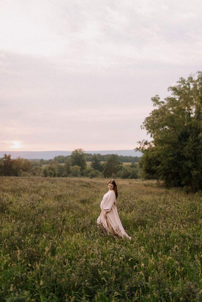 Pregnant woman in beige dress cradling baby bump in open field at sunset