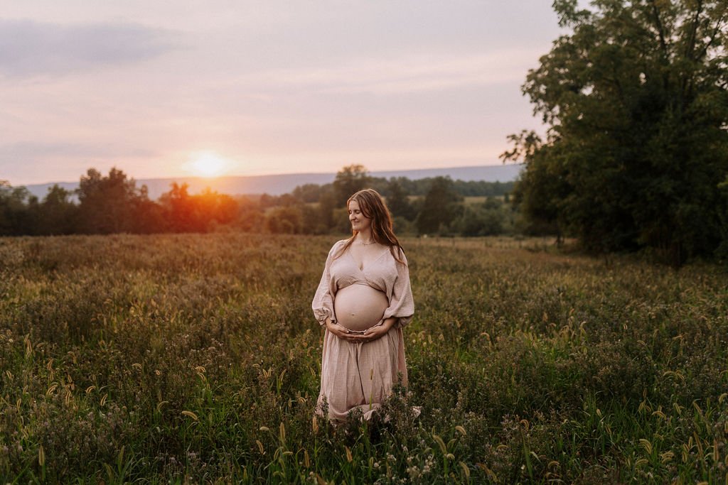 Pregnant woman in flowing beige dress standing in grassy field with trees in background