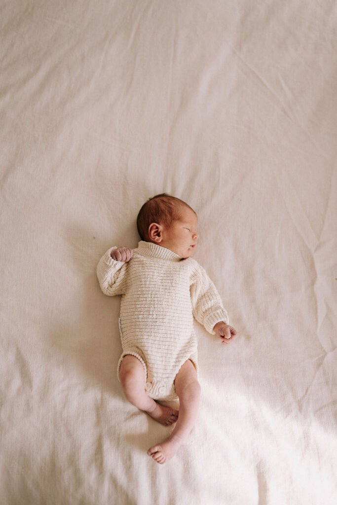 Newborn baby in cream knit outfit lying on white bedding