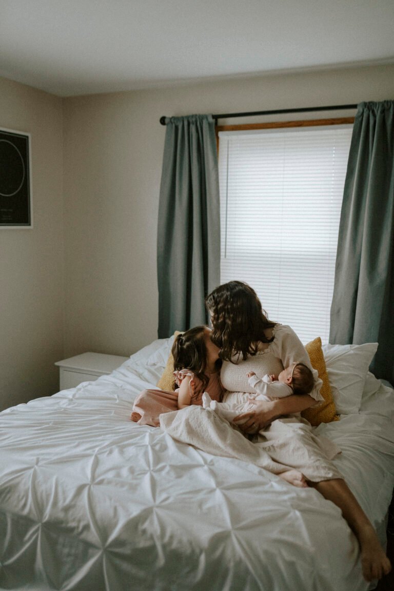 Mom holding newborn daughter sitting with big sister on bed during newborn pictures