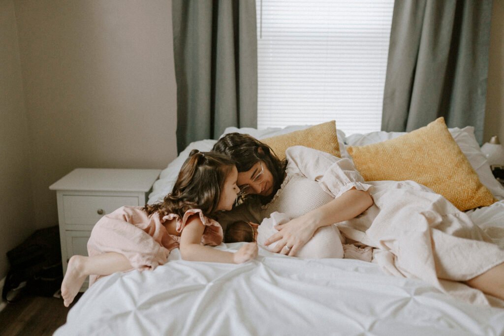 mom in cream dress cuddling with newborn on bed while sister is standing over them